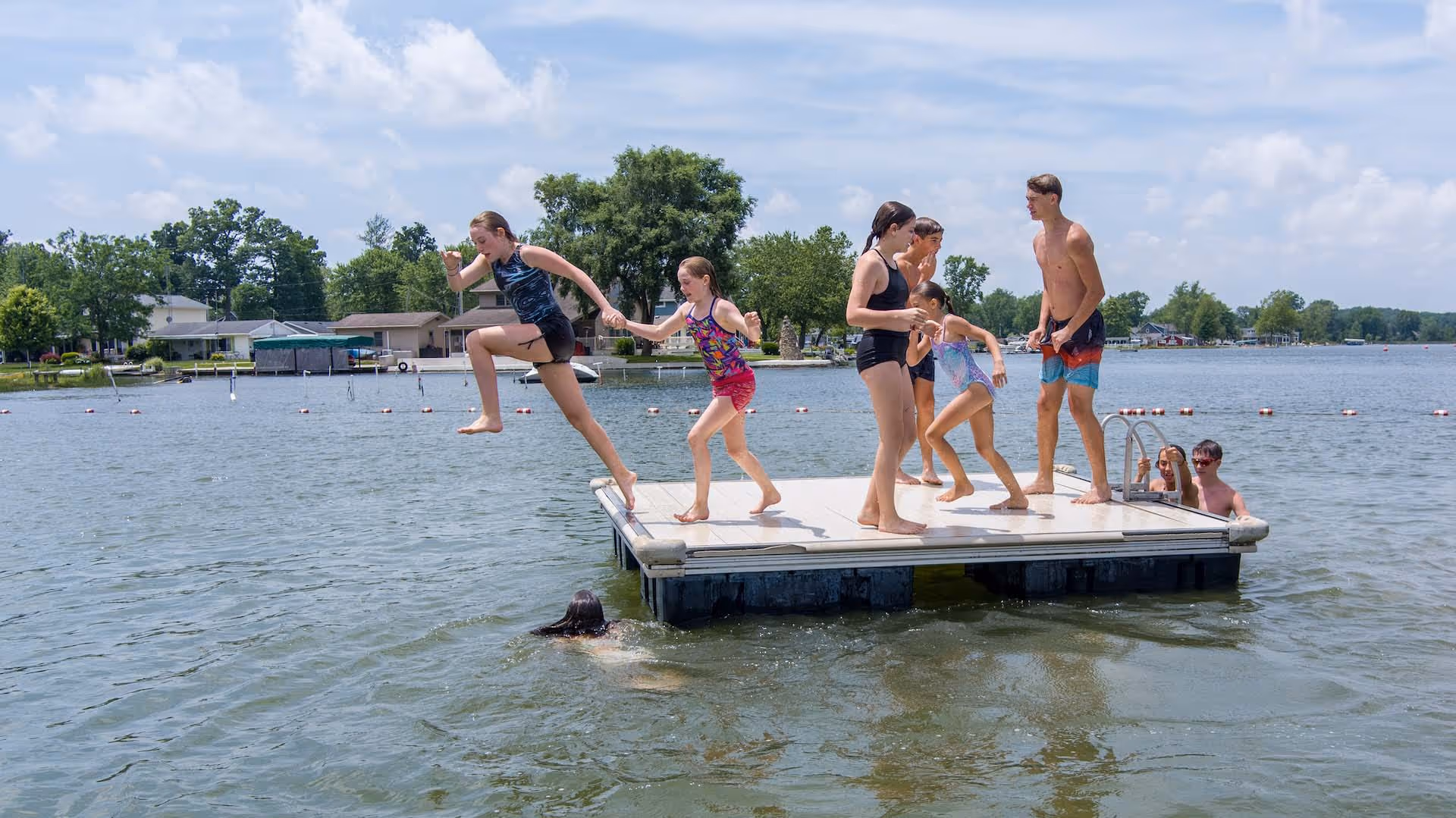 Children and teenagers playing and jumping off a floating dock into a lake on a sunny day.