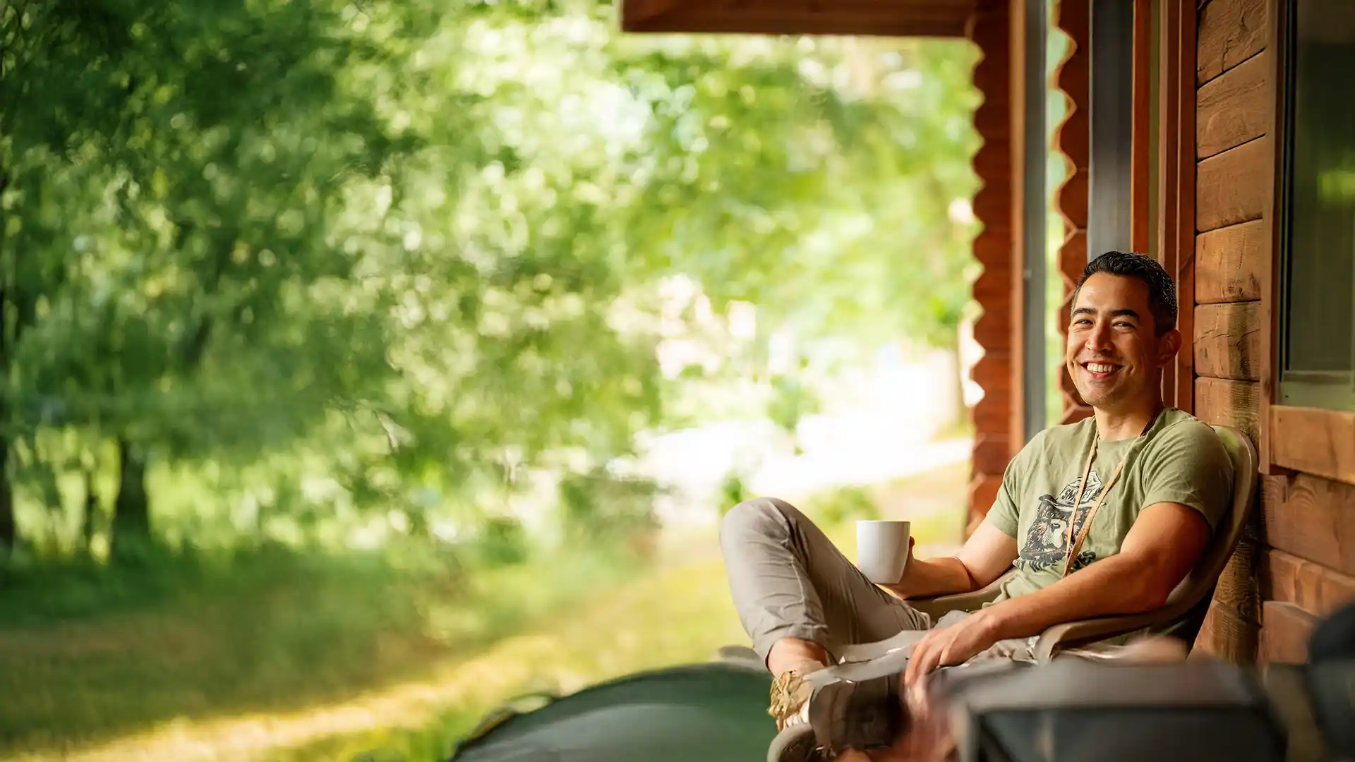 Smiling man in casual clothes sitting on a porch chair holding a white mug with greenery in the background.