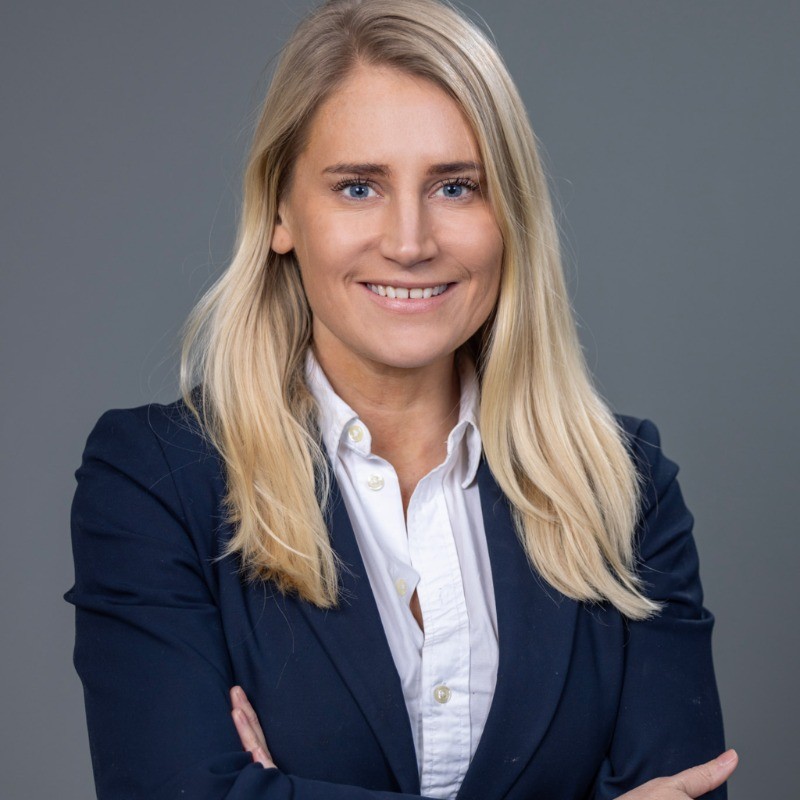 Porträtt av leende kvinna med långt blont hår i vit skjorta och mörk blazer mot grå bakgrund.