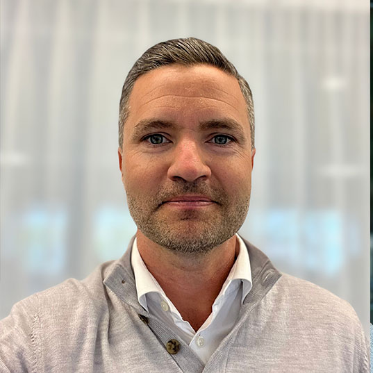 Man wearing a red First Camp sweatshirt sitting inside a camper with a window showing caravans outside.