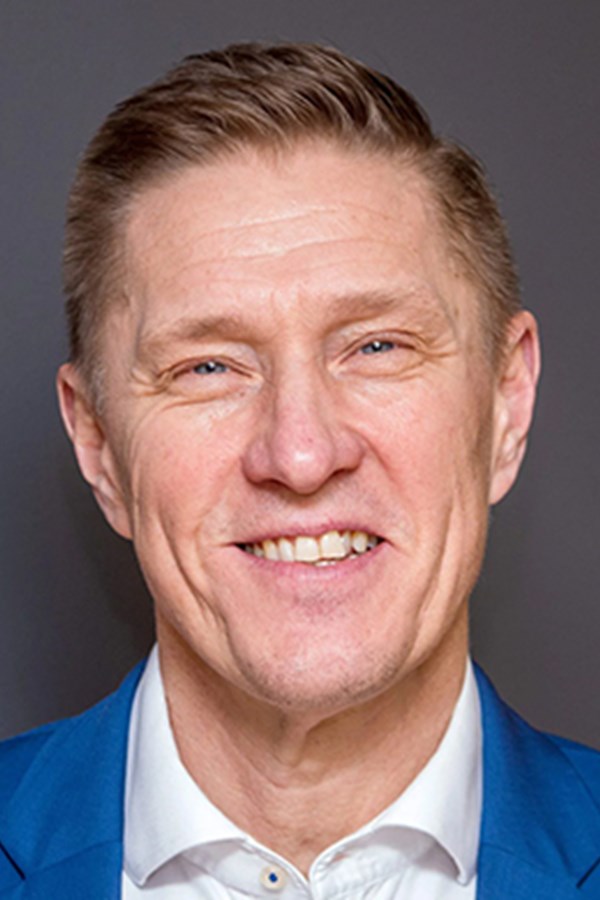 Man with short dark hair, wearing glasses and a blue shirt, smiling with green foliage in the background.