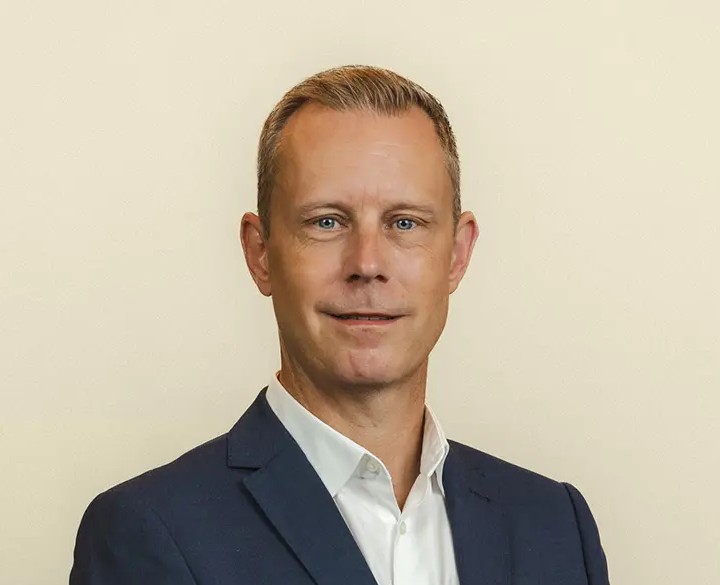 Porträtt av leende kvinna med långt blont hår i vit skjorta och mörk blazer mot grå bakgrund.