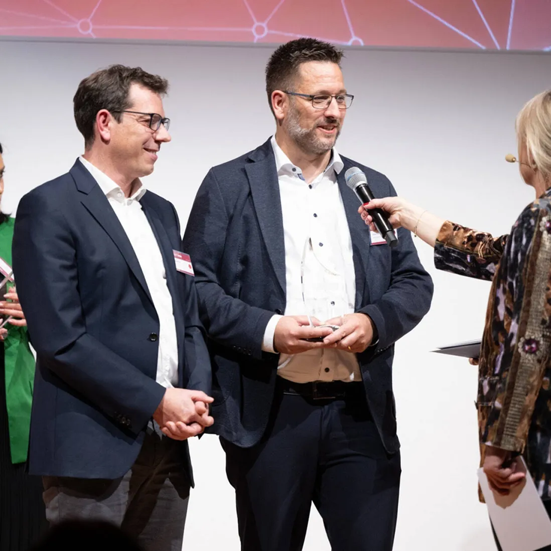 Two men in suits, one holding a clear award, being interviewed with a microphone by a woman on stage.