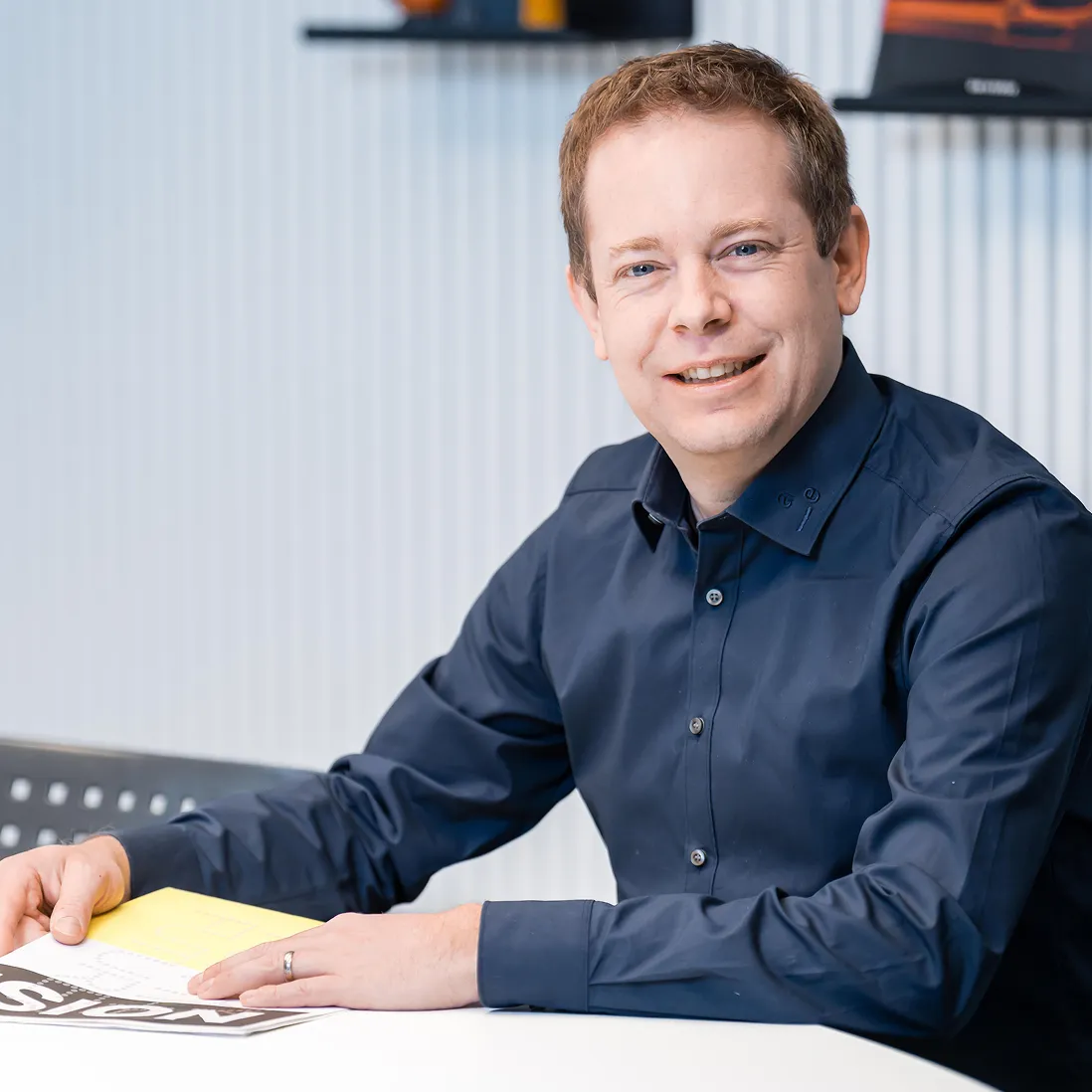Smiling man in a dark blue shirt sitting at a table with a magazine or document.