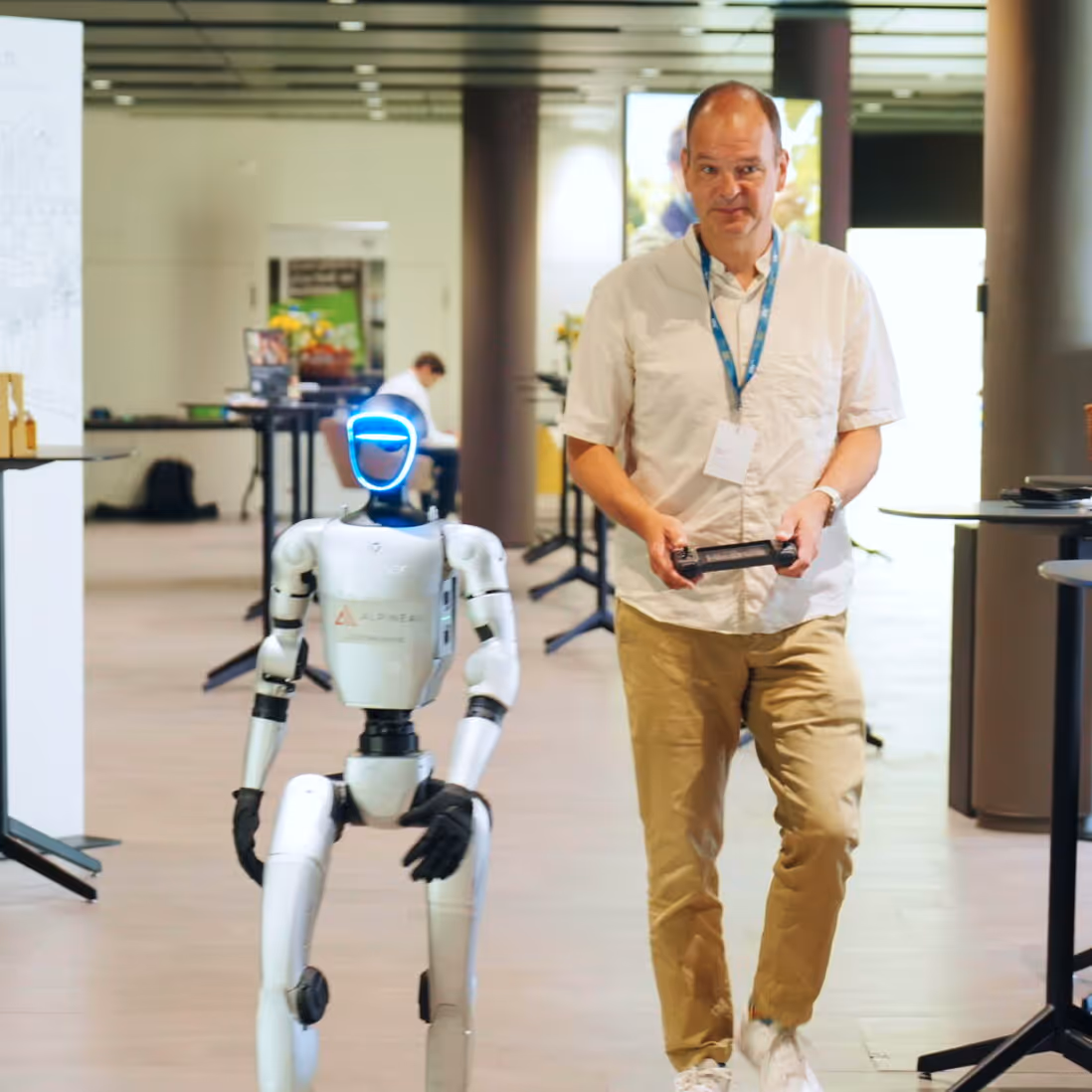 Two men sitting across from each other at a table, one wearing a virtual reality headset and the other with hands near face in discussion.