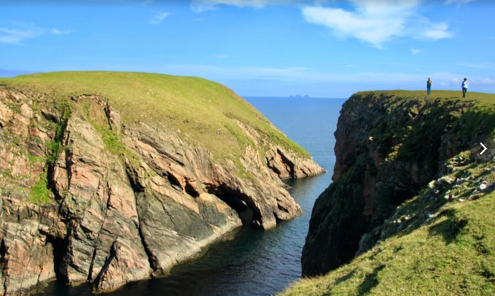 Grassy cliffs overlooking a narrow ocean inlet under a blue sky with two people standing on the right cliff.