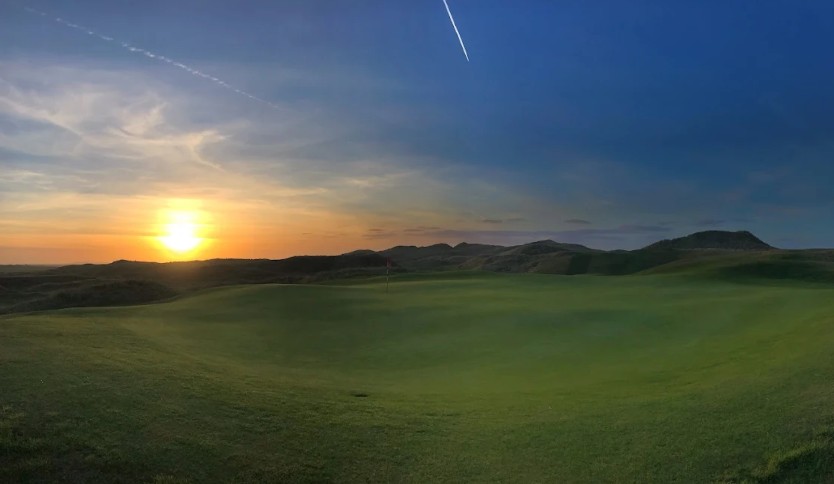 Golf course green with flag set against a sunset and a partly cloudy sky with airplane contrails.