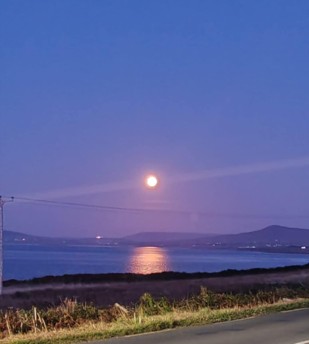 Full moon reflecting on calm water with hills in the background and a road in the foreground at dusk.
