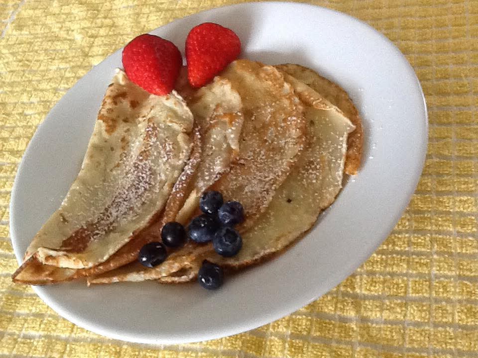 White plate with four folded crepes dusted with powdered sugar, topped with two strawberries and several blueberries, placed on a yellow textured cloth.