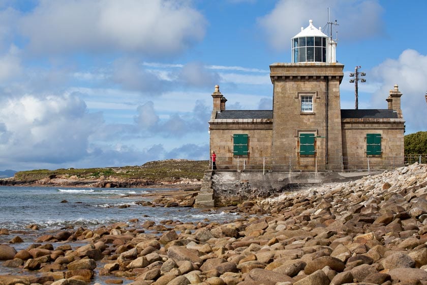 Stone lighthouse with green shuttered windows by a rocky shore under a partly cloudy sky.