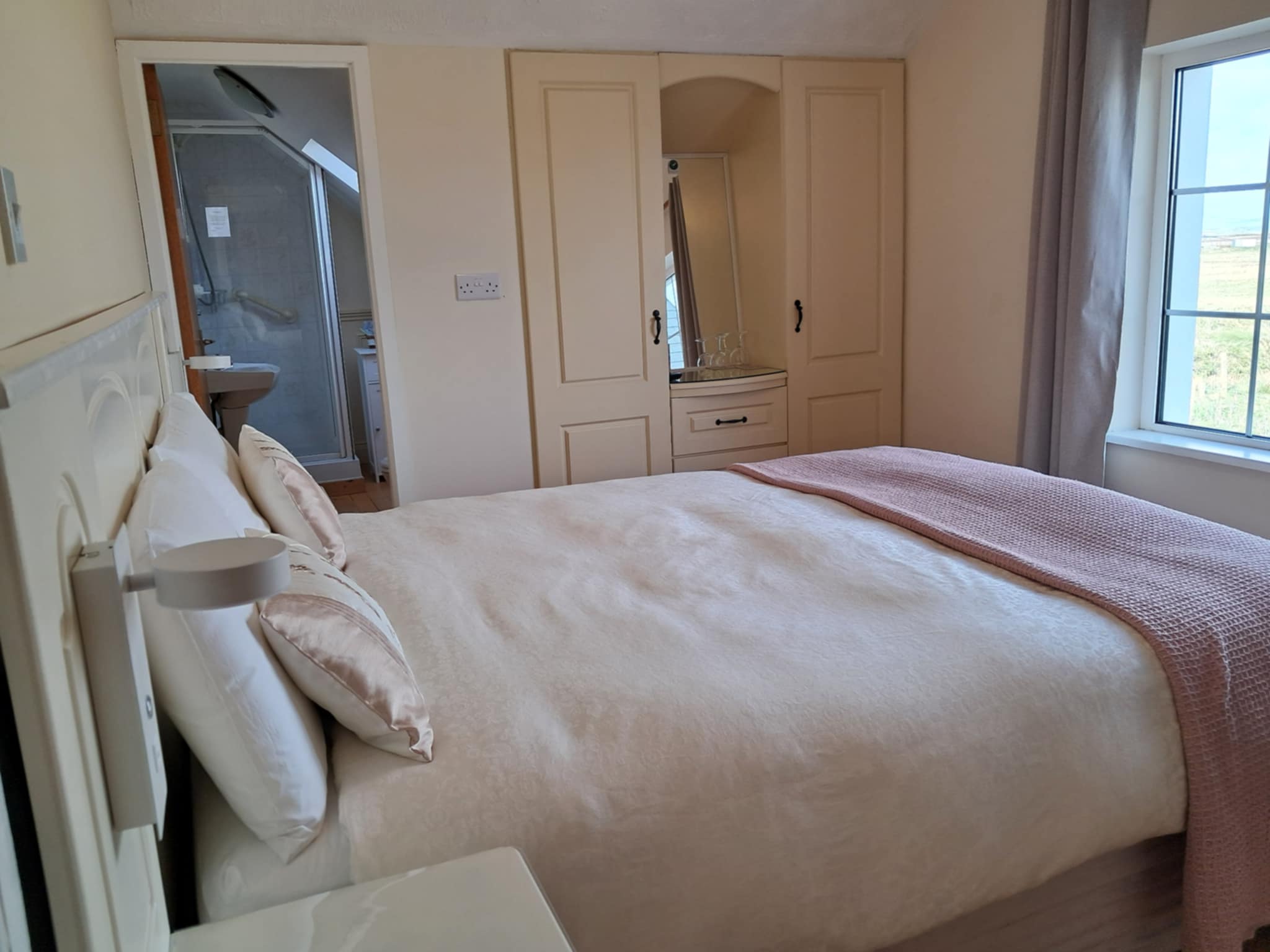 Cozy bedroom with a cream-colored bedspread, pale pink blanket, pillows, a built-in closet with a mirror, and a view of the countryside through a large window.