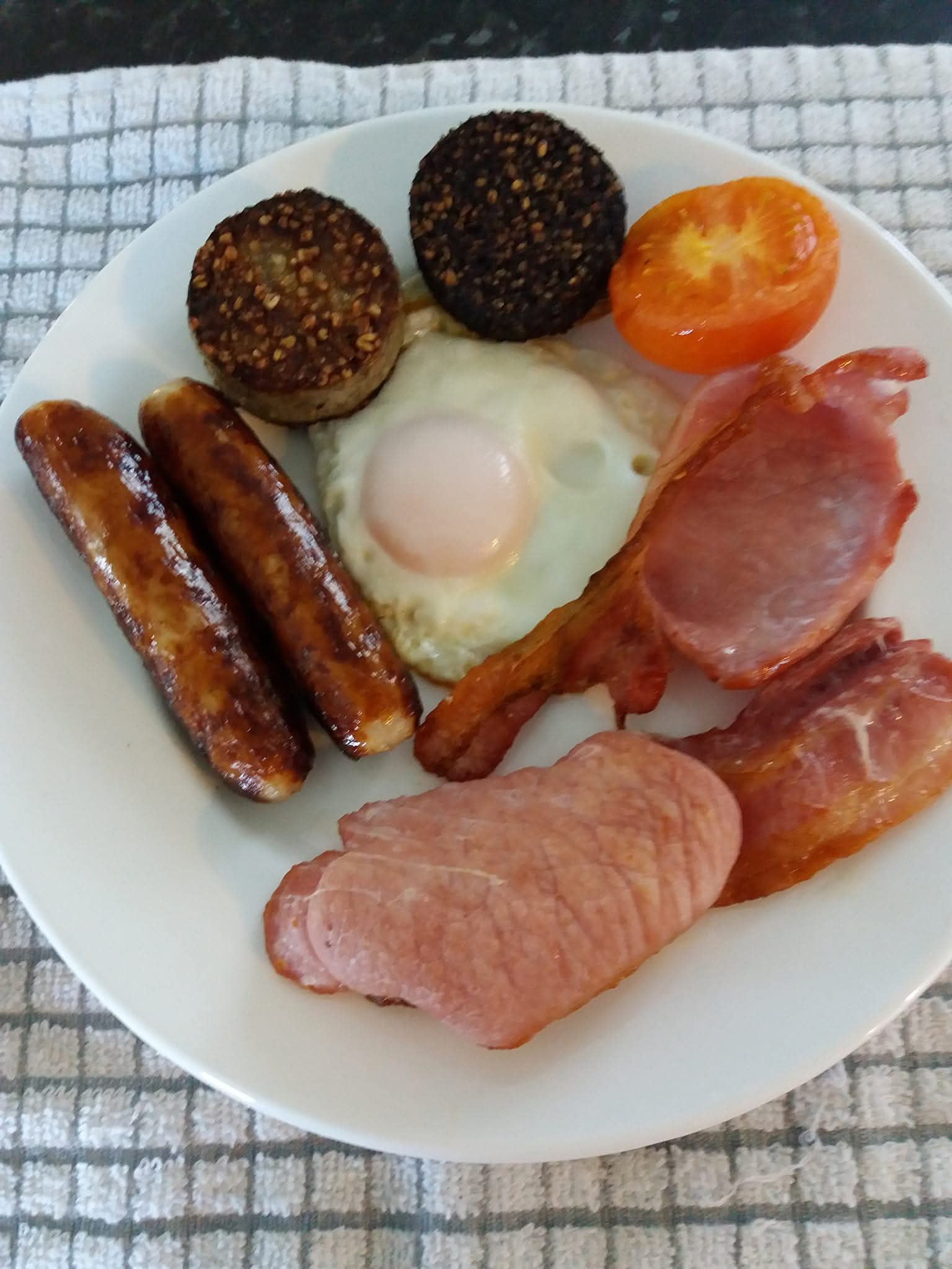 Plate with cooked breakfast including two sausages, black pudding, fried egg, grilled tomato half, two strips of bacon, and a slice of ham.