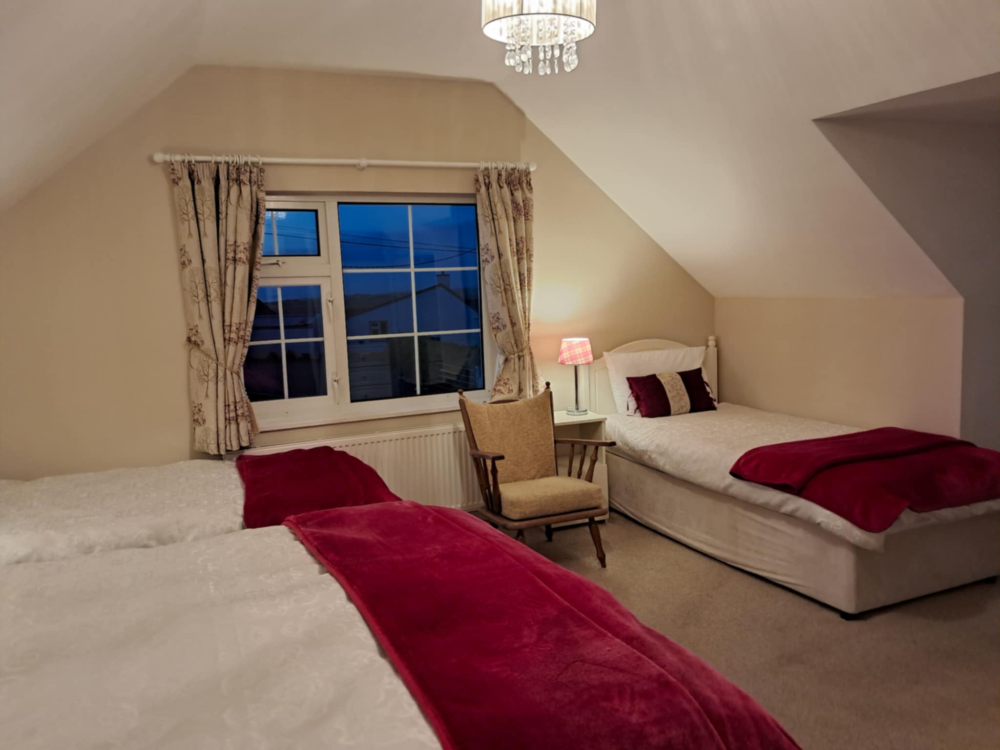 Cozy bedroom with two single beds dressed in white linens and red throws, a padded wooden armchair, and a window with floral curtains at dusk.