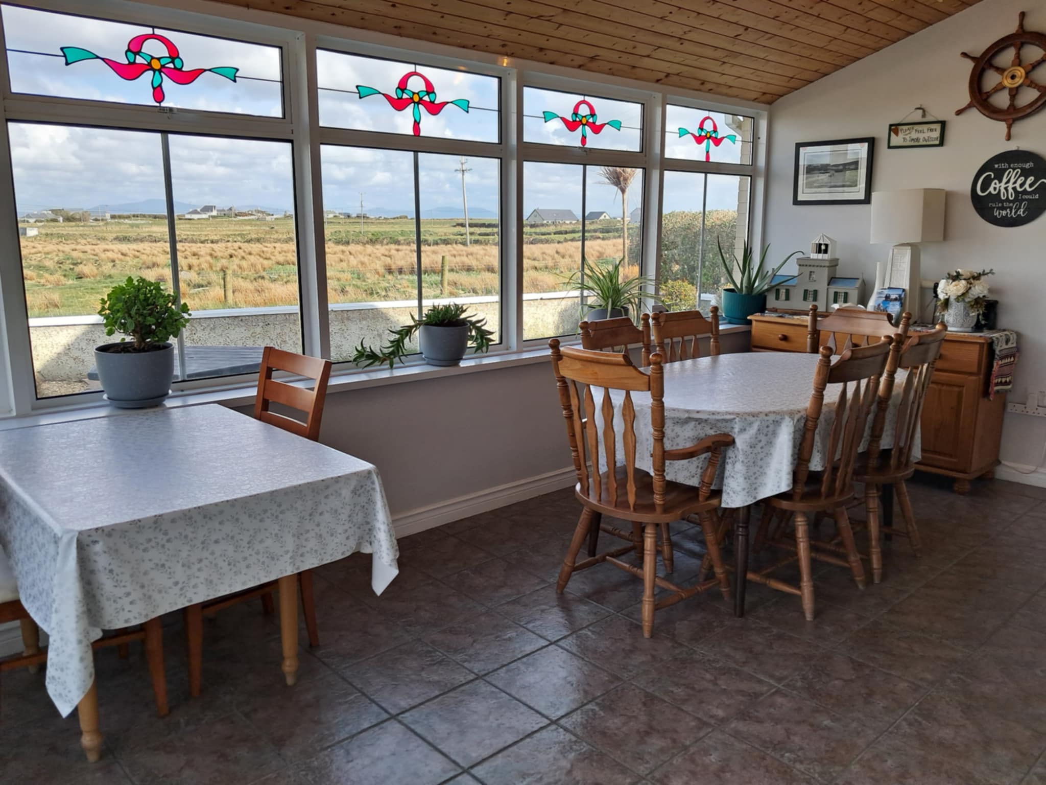 Cozy dining area with two tables covered in light floral tablecloths, wooden chairs, potted plants on the windowsill, and large windows featuring decorative red and turquoise stained glass designs.