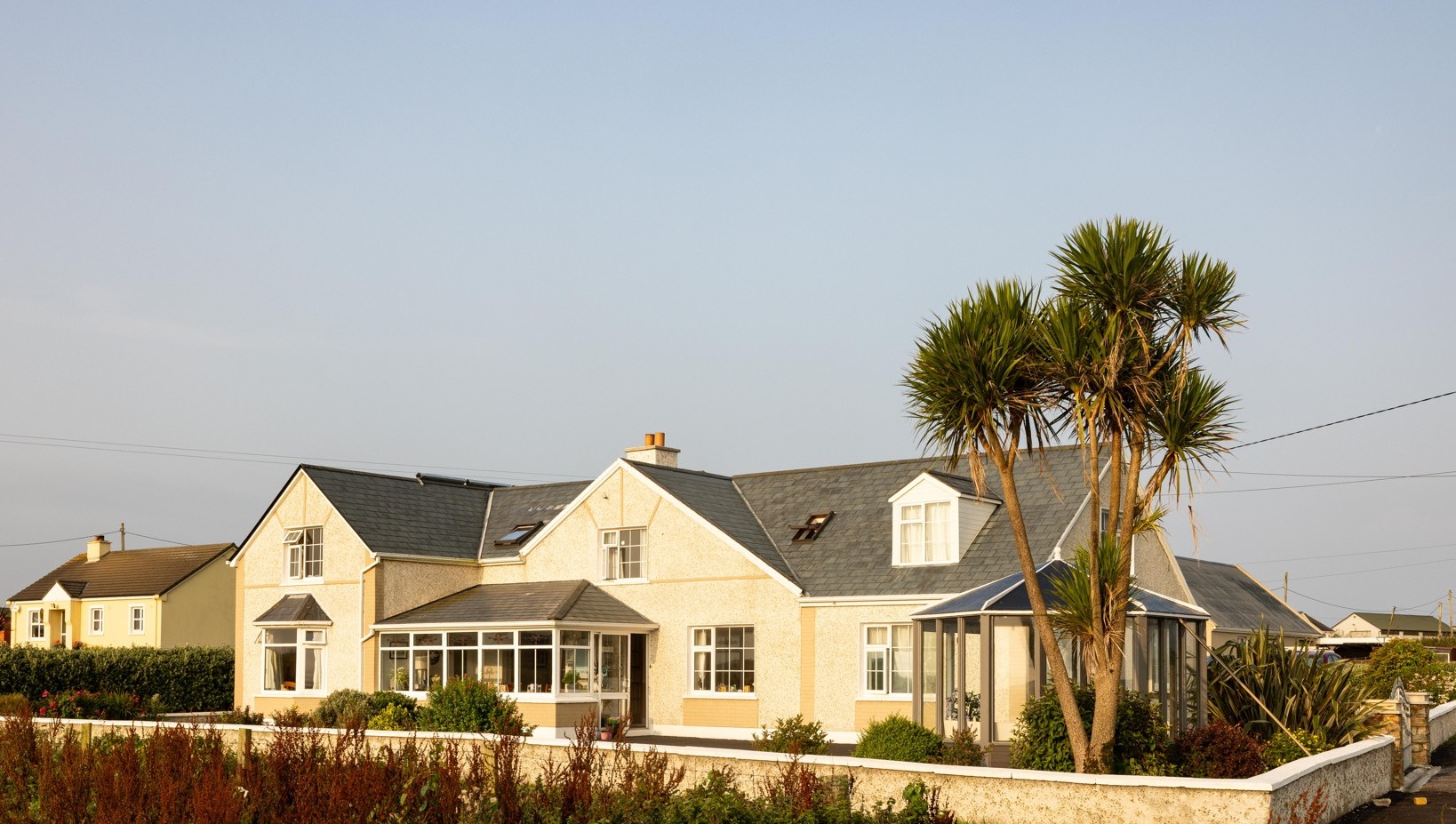 Cream-colored residential house with gray roof, sunroom, and tall palm trees in the front yard under a clear sky.