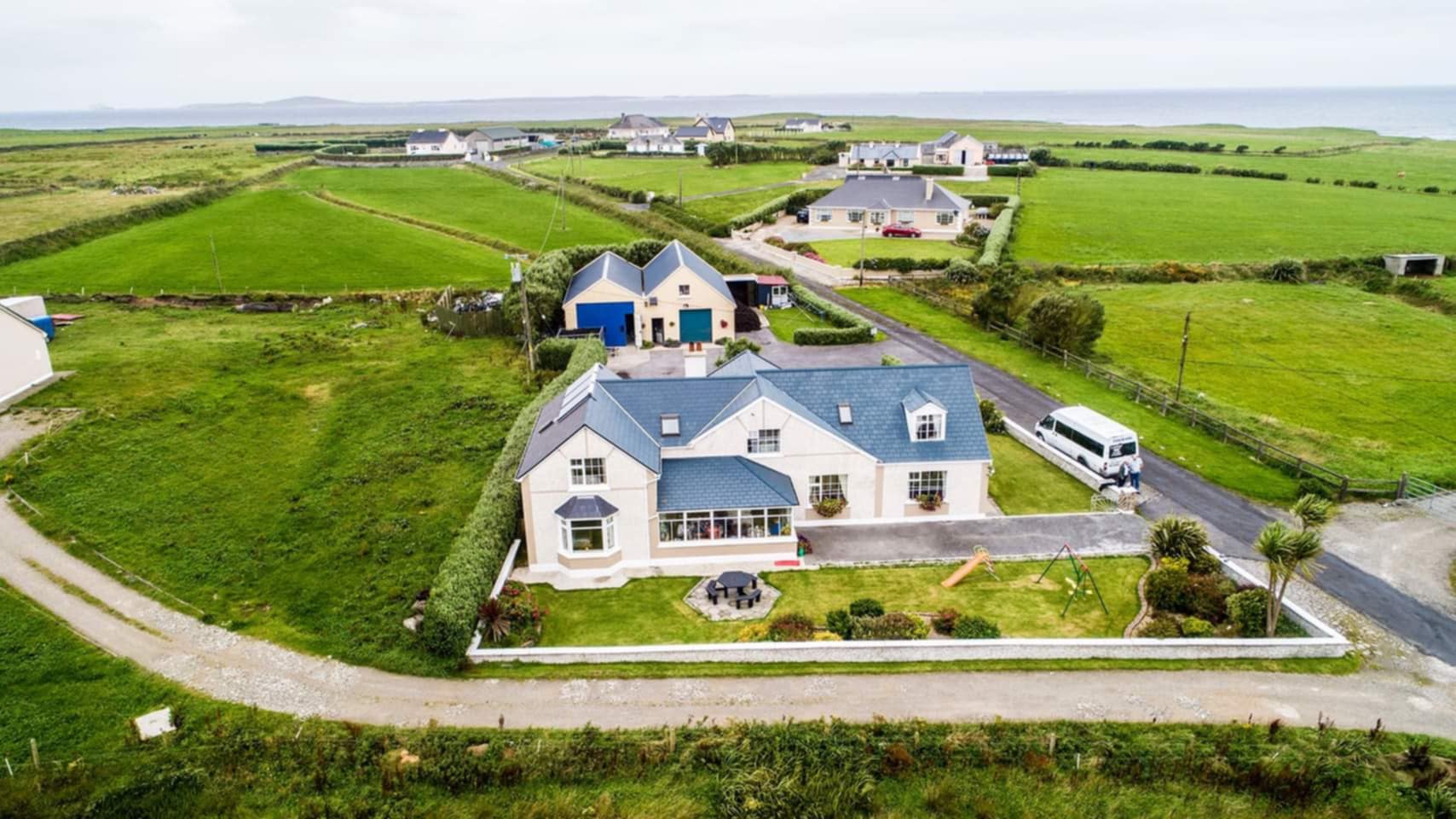 A large house with blue-gray roofing surrounded by green lawns, gardens, and a playground in a rural area with other houses and fields extending to the horizon.