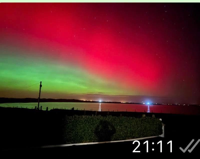 Night sky showing vibrant red and green northern lights above a quiet body of water with distant lights on the horizon.