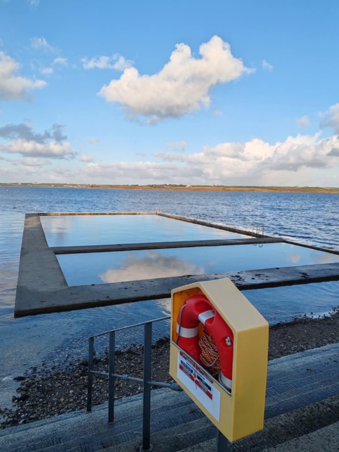 Outdoor rectangular tidal swimming pool beside a body of water with a life preserver mounted on a yellow box in the foreground.