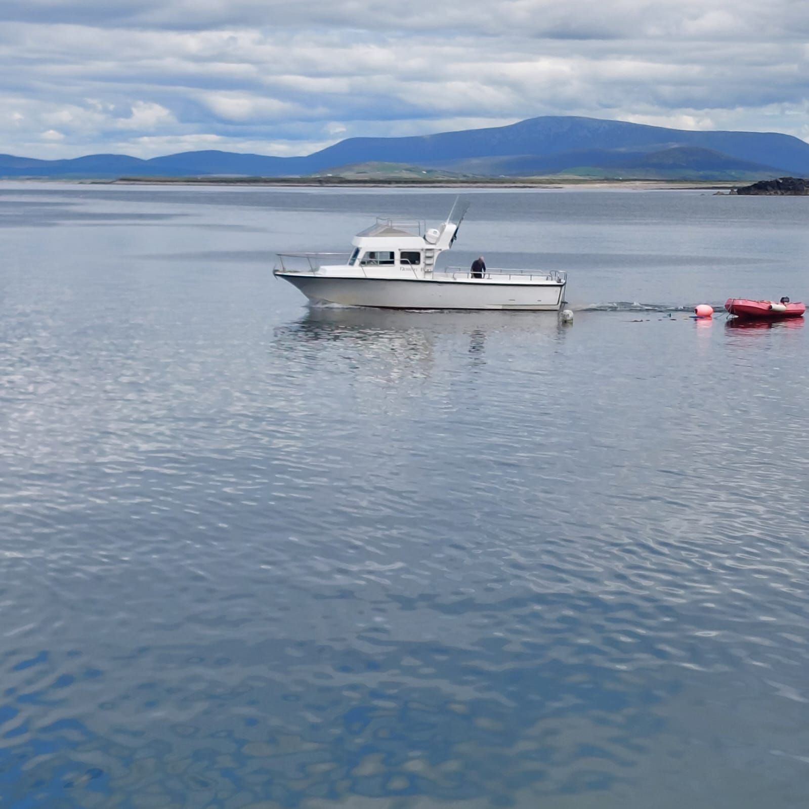 White motorboat anchored on calm water with a person on board, distant shoreline and blue mountains under a cloudy sky.