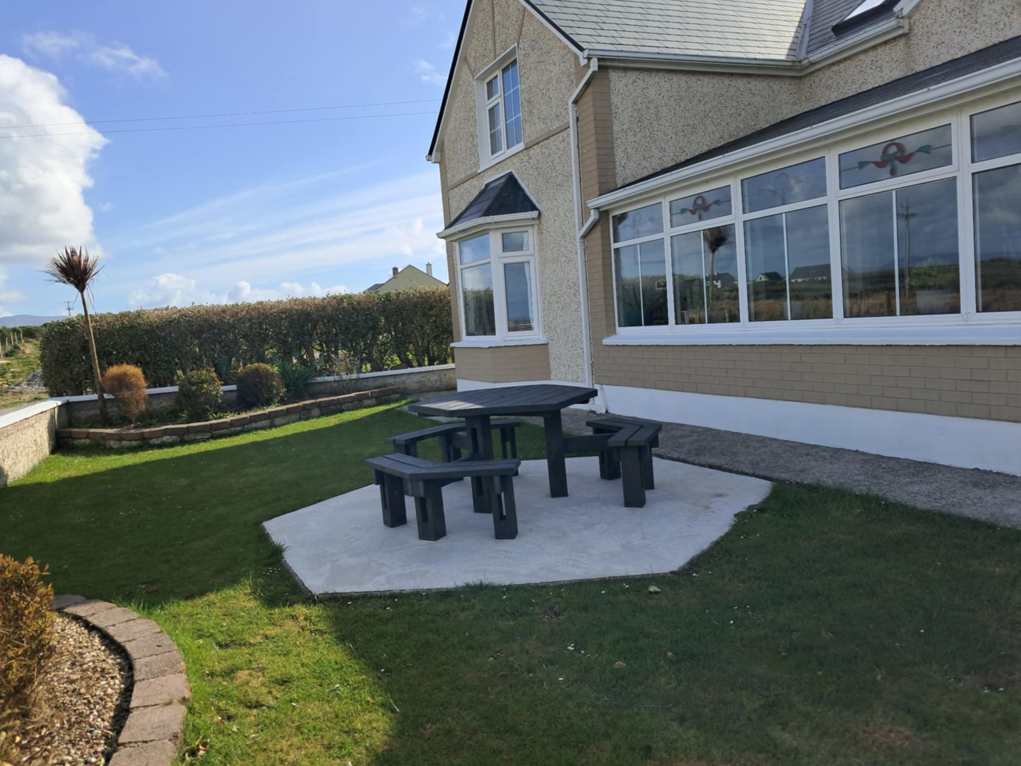 Black octagonal outdoor picnic table with benches on a concrete patio beside a beige house with large windows and green lawn.