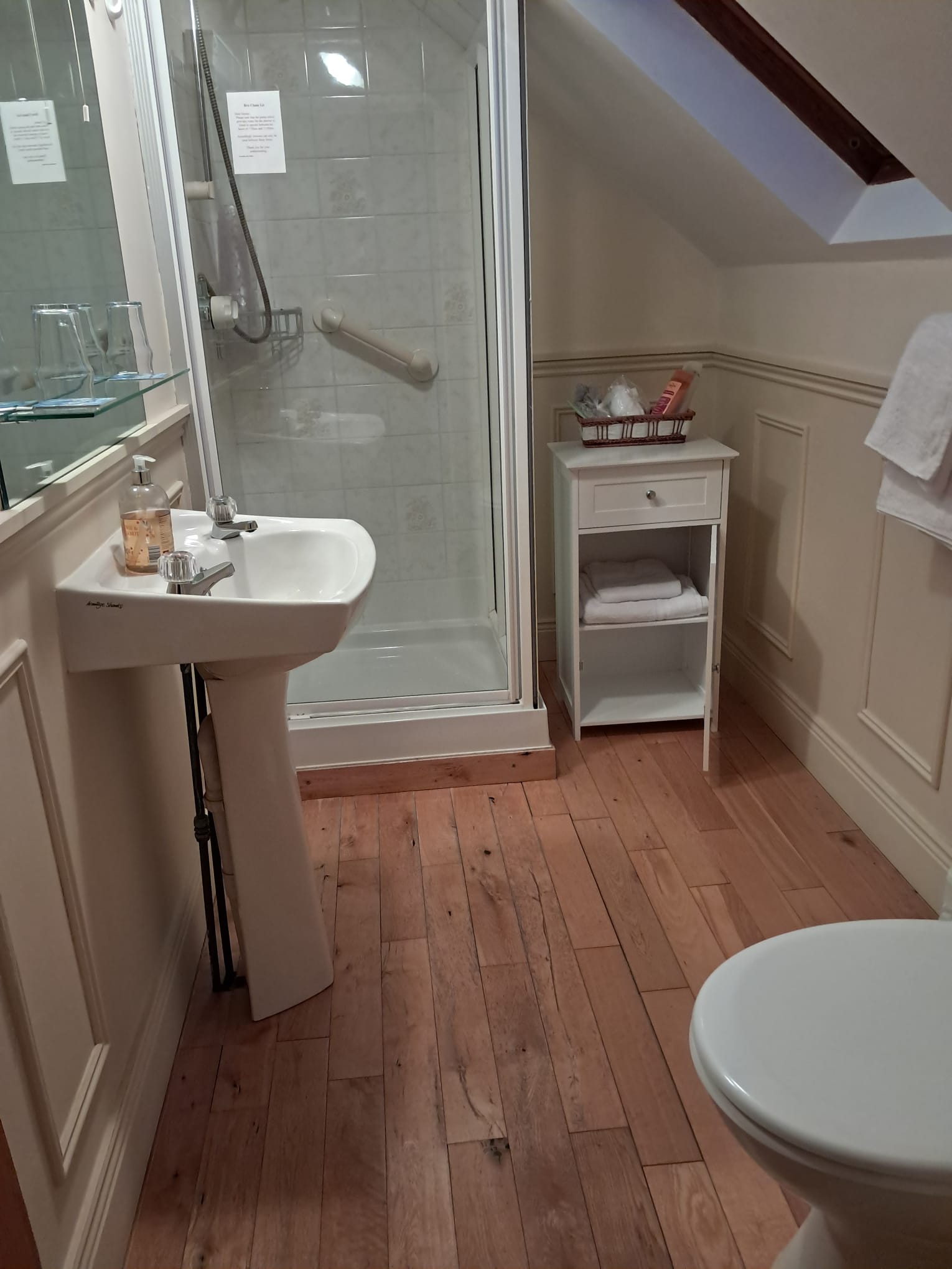 Compact bathroom with wooden floor, white pedestal sink, glass-enclosed shower, small white cabinet with towels, and toilet.