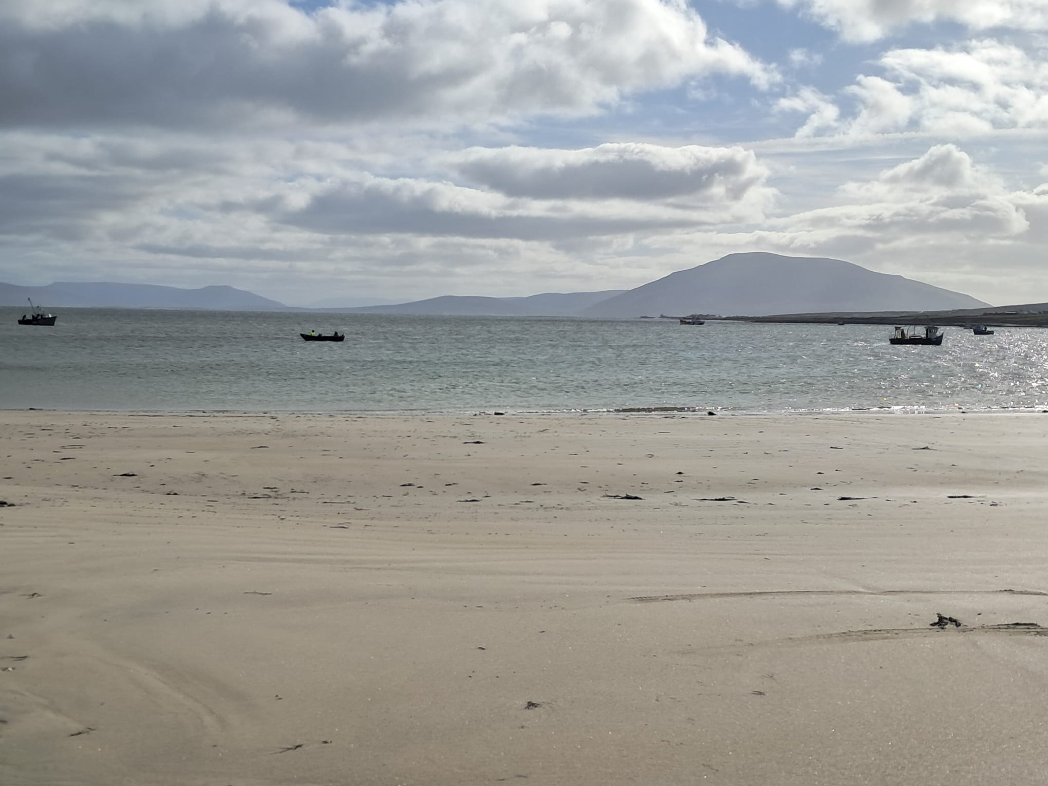 Sandy beach with calm sea, small fishing boats on the water, and hills under a partly cloudy sky.