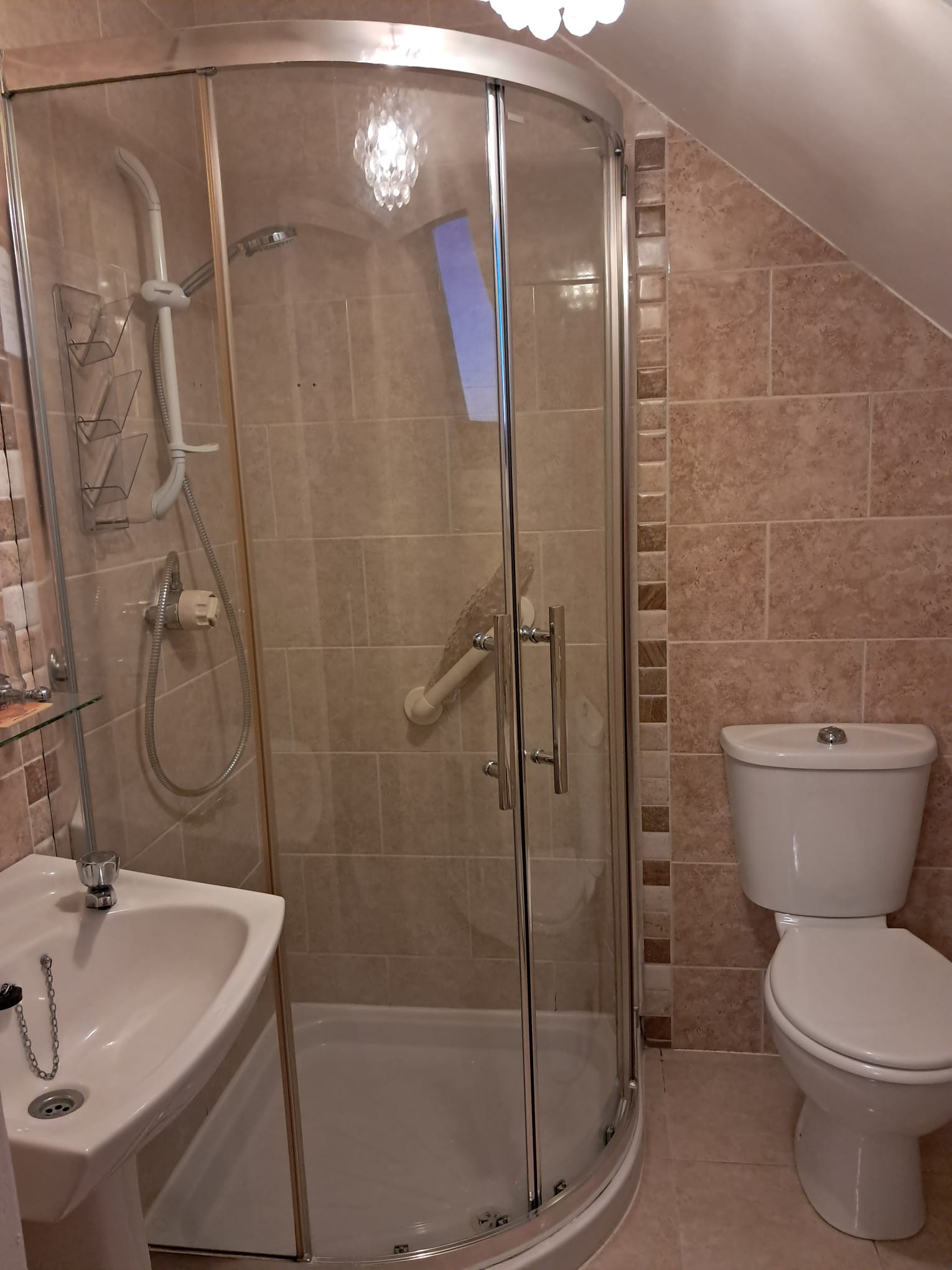 Small bathroom with a corner glass shower enclosure, a white toilet, and a white pedestal sink, all set against beige tiled walls.