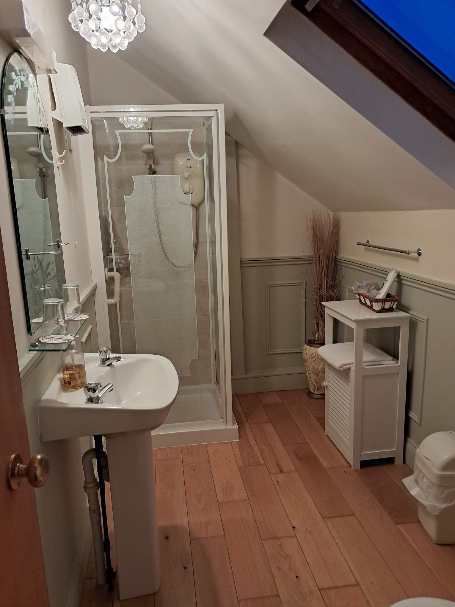 Bathroom with wooden floor, white pedestal sink, glass-enclosed shower, small white cabinet with towels, and sloped ceiling with skylight.