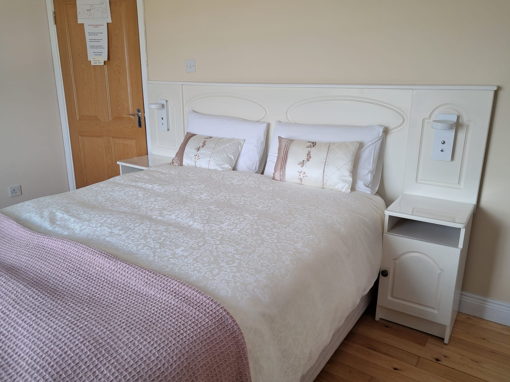 Neatly made double bed with white floral bedspread, two decorative pillows, white headboard, and bedside tables in a room with wooden floor.
