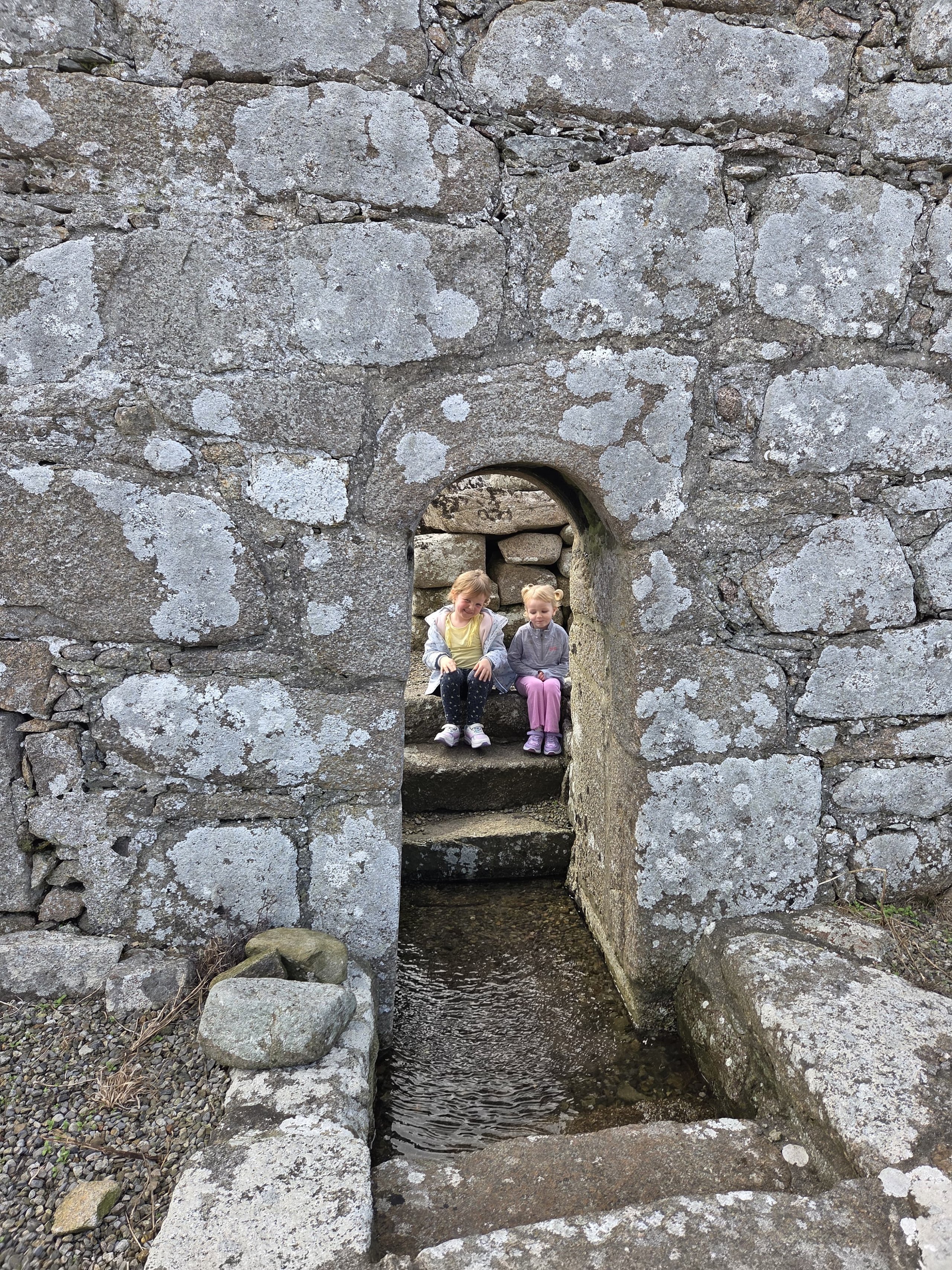 Two young girls sitting on stone steps inside a moss-covered stone archway over a narrow water channel.