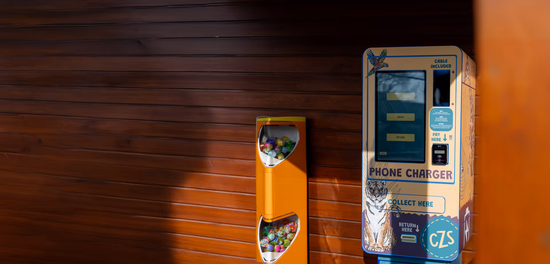 Phone charger vending machine with illustrations of a tiger and a parrot, next to an orange capsule toy dispenser against a wooden wall.