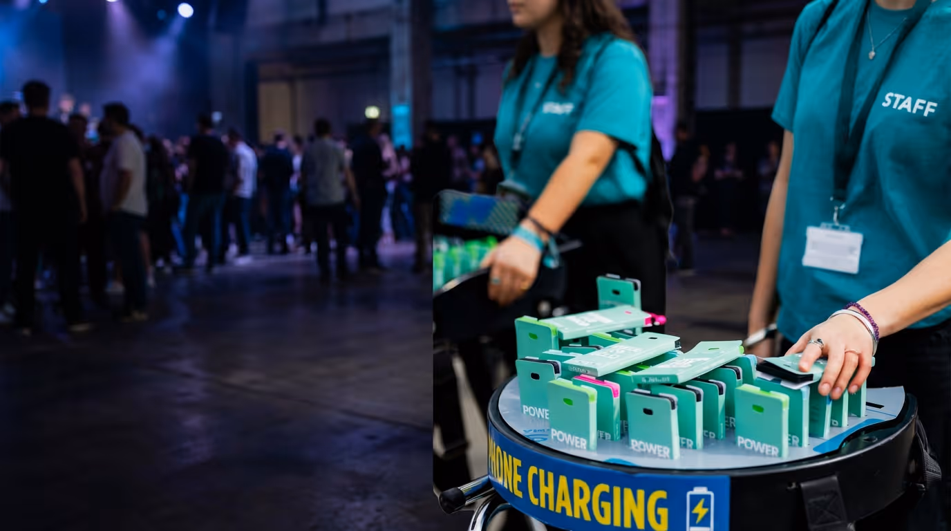 Staff members in teal shirts managing a phone charging station with green power banks in a crowded event space.
