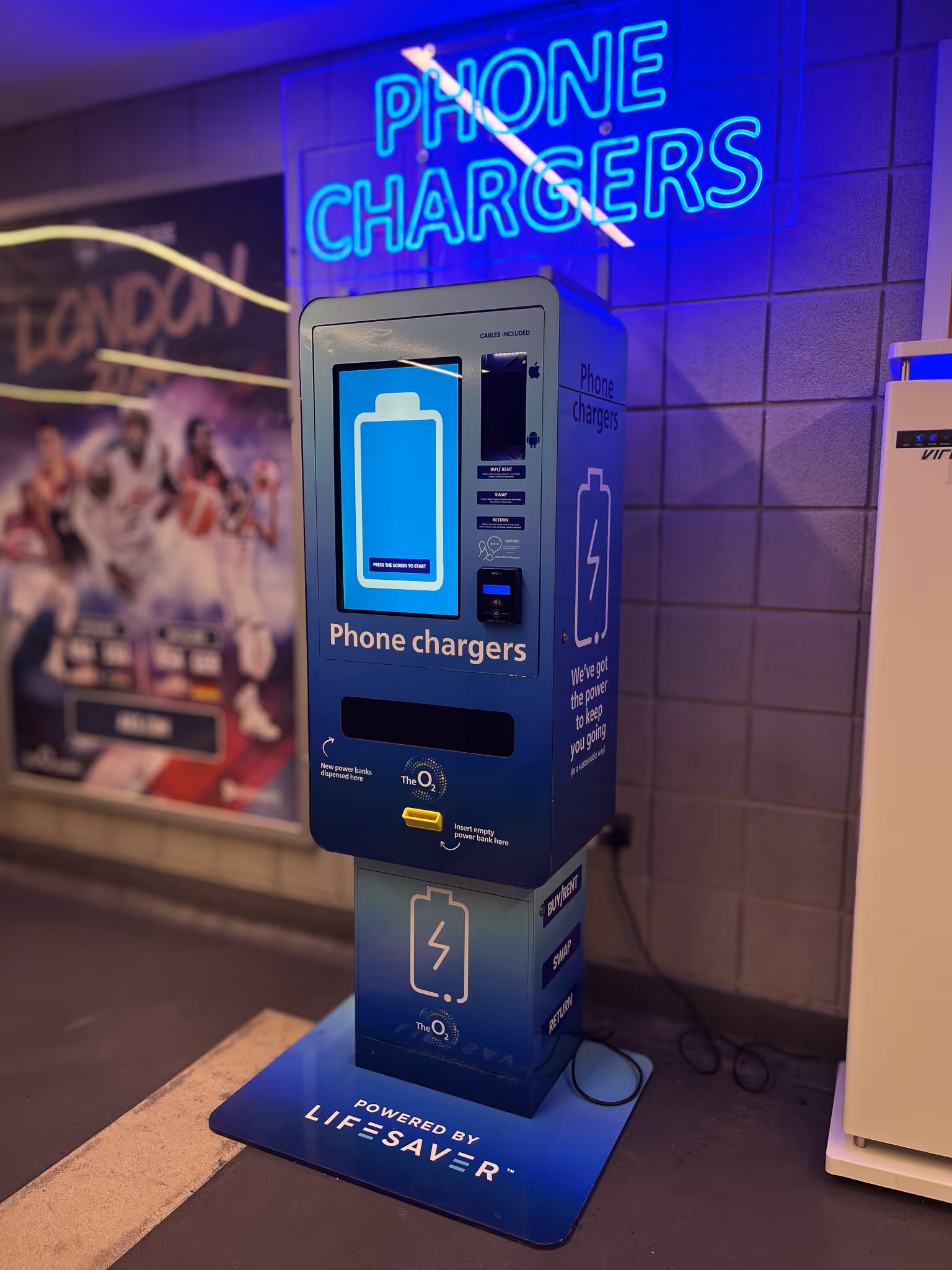 Blue phone charger rental kiosk with touchscreen and neon 'PHONE CHARGERS' sign above, placed indoors near a wall.
