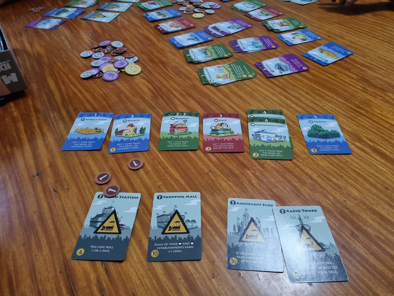 Gameplay photo of the board game Machi Koro featuring various components and board state.