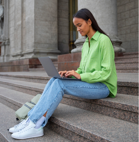 Young woman in a green shirt sitting on outdoor stone steps working on a laptop with a backpack beside her.
