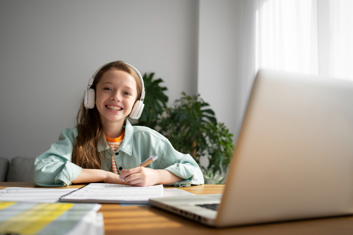 Smiling girl wearing headphones writing in a notebook during online learning at a wooden table with a laptop.