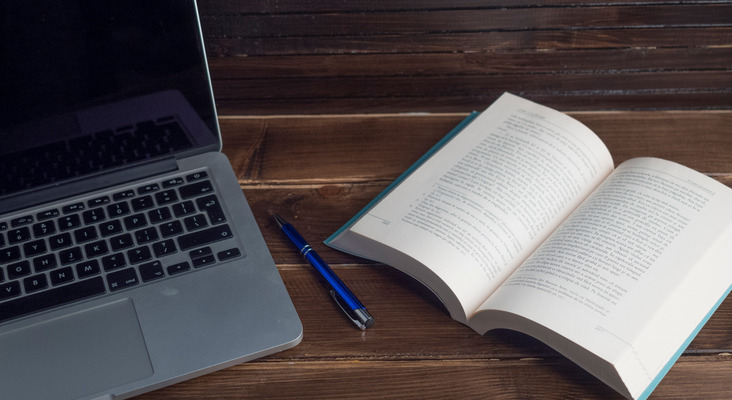 Open book, blue pen, and laptop on a wooden surface.