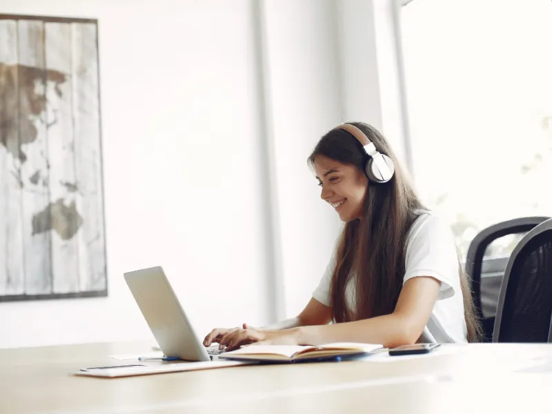 Young woman wearing headphones working on a laptop at a desk with an open notebook and smartphone.