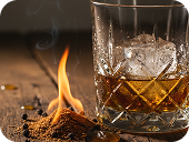 Glass of whiskey with ice on wooden surface next to a small flame and scattered peppercorns.