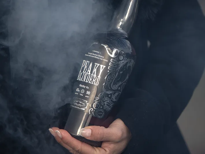 Hand holding a dark bottle labeled 'Peaky Blinders Shabby Co.' partially obscured by smoke.