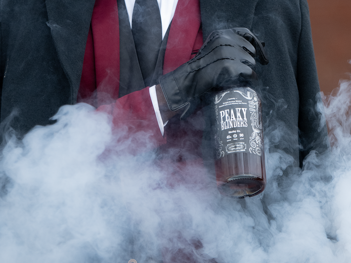 Person wearing black gloves and a red suit holding a smoking bottle labeled 'Peaky Blinders'.