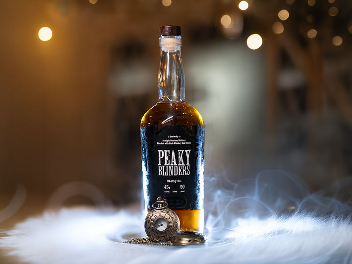 Bottle of Peaky Blinders bourbon whiskey with a vintage pocket watch on a misty surface under warm ambient lights.
