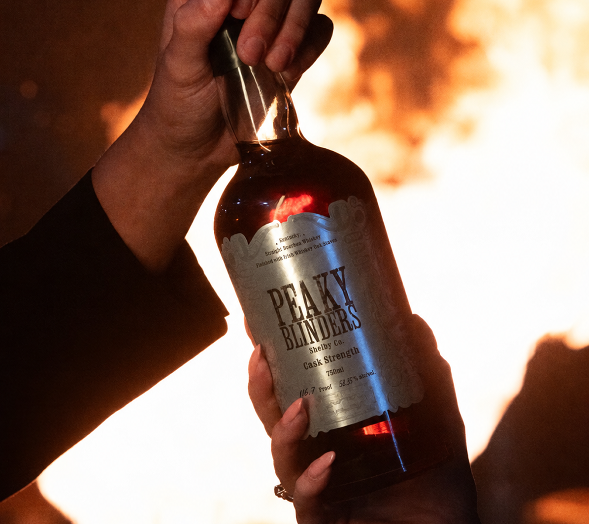 Two hands holding a bottle of Peaky Blinders Shelby Co. Cask Strength whiskey against a fiery background.