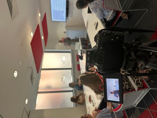 Photograph of a professional training seminar being filmed, with a camera in the foreground recording a speaker presenting to attendees in a modern meeting room.