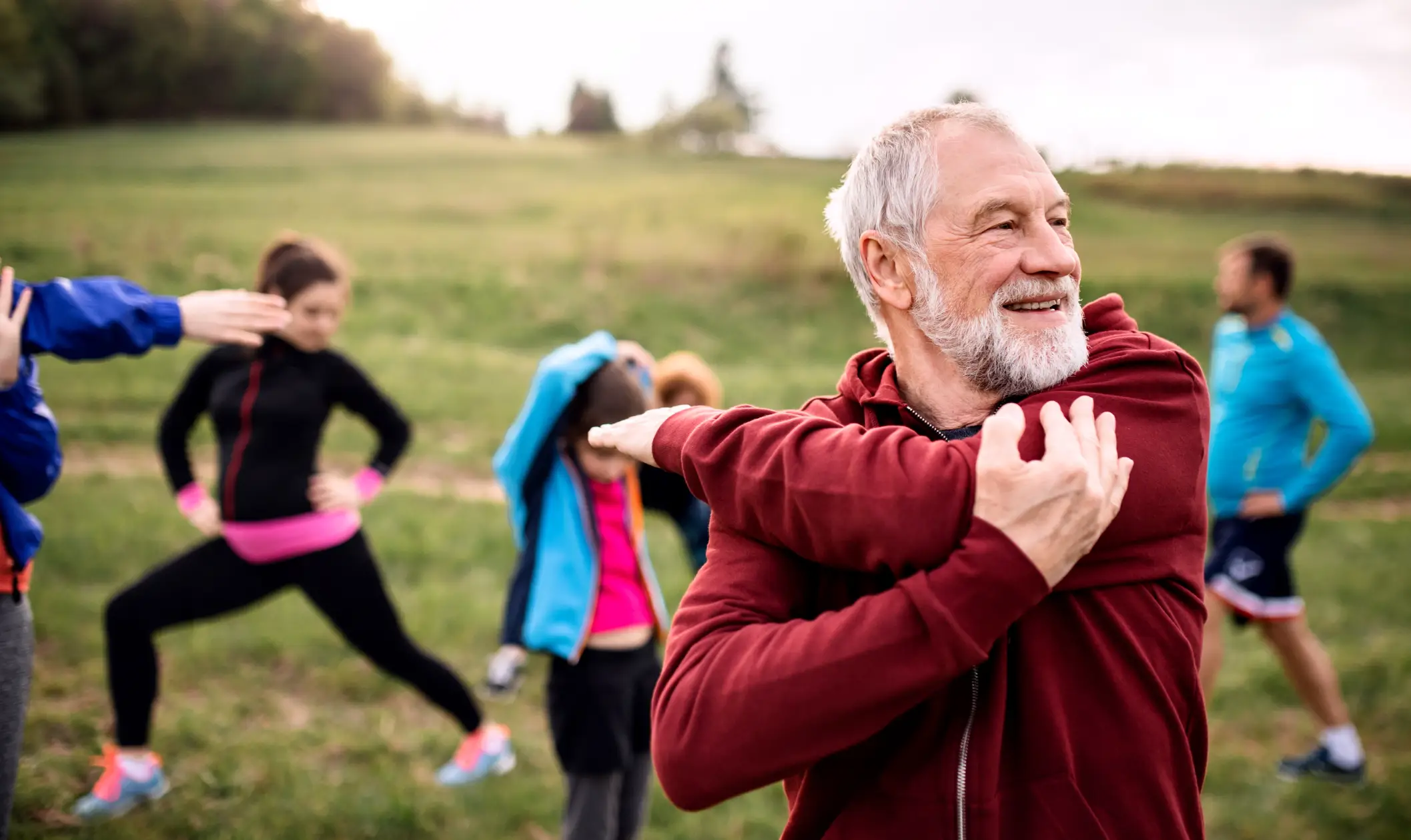An older man outdoors in exercise attire, stretching his arm across his body with a smile, showing an active lifestyle.