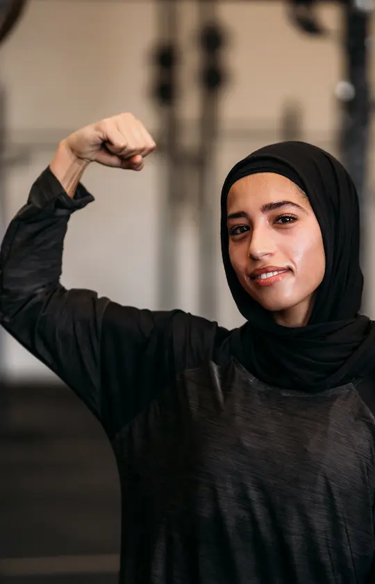 Woman in a black hijab and sportswear flexing her arm muscle with a confident smile.