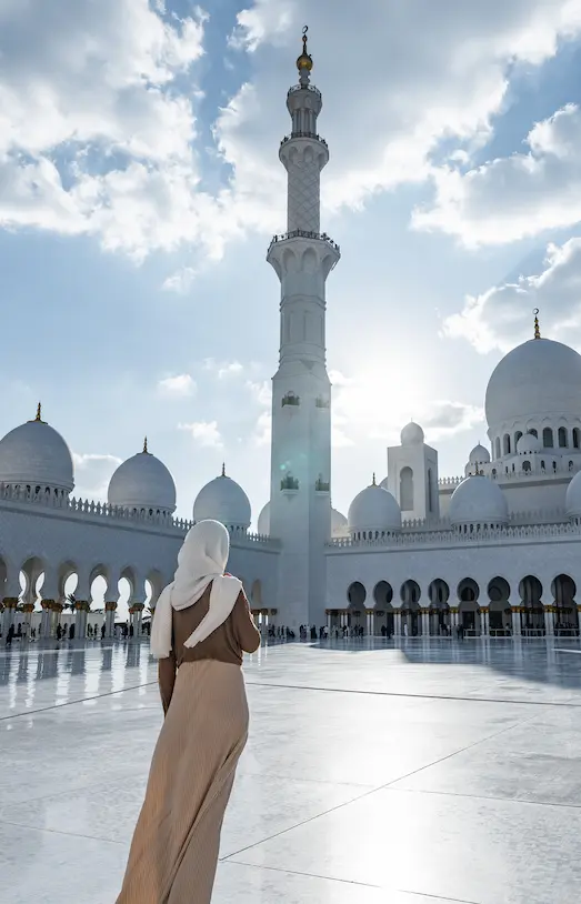 Woman wearing a headscarf standing in the courtyard of Sheikh Zayed Grand Mosque with white domes and minarets under a partly cloudy sky.