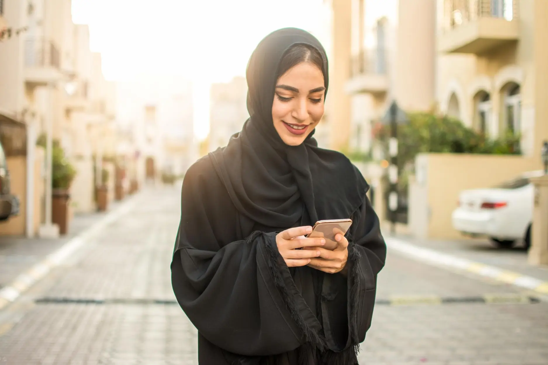 Smiling woman in black hijab standing on a sunlit street using a smartphone.