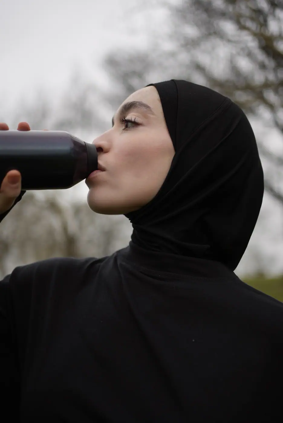 Young woman wearing a black hijab and shirt drinking from a black water bottle outdoors.