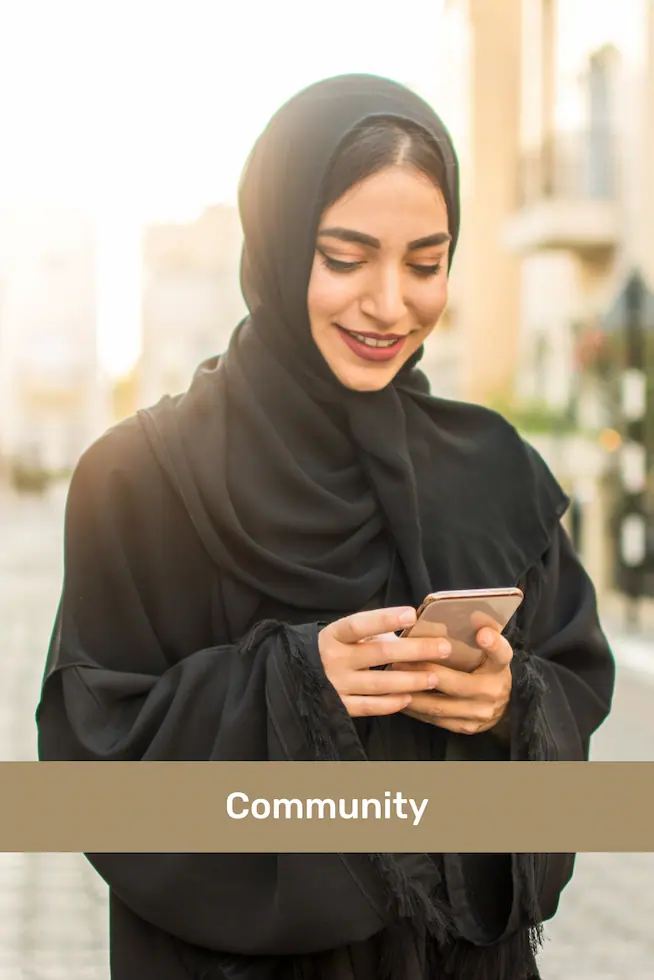 Woman wearing a black hijab looking at her phone and smiling outdoors with buildings in the background.
